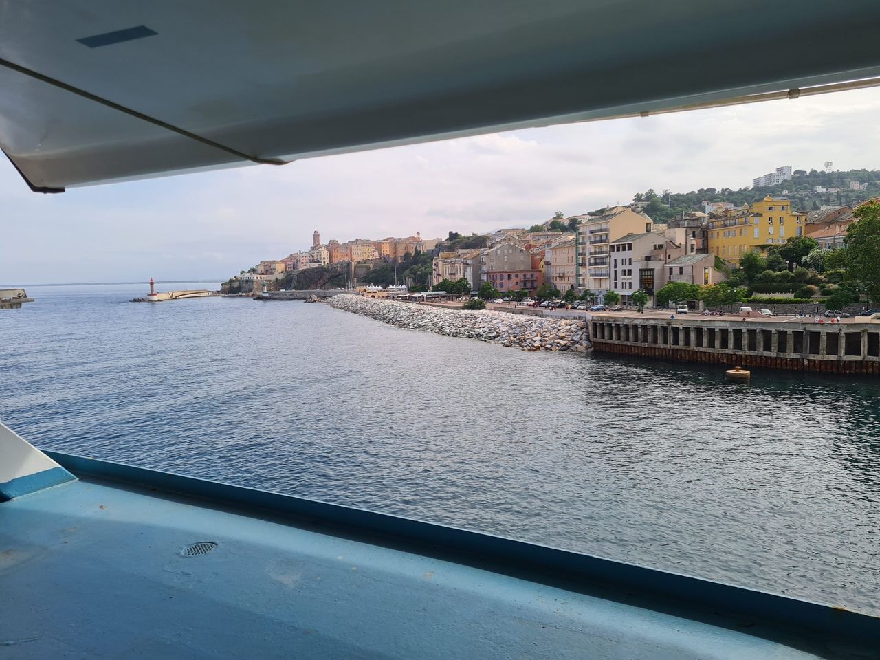 View from a boat beneath its wing toward a calm harbor, rocky shoreline, colorful waterfront buildings, and hillside homes under a cloudy sky.