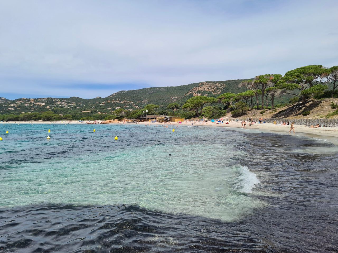 Clear turquoise bay with yellow buoys, a sandy beach, umbrella pines, and green hills under a cloudy sky; peaceful coastal scene.