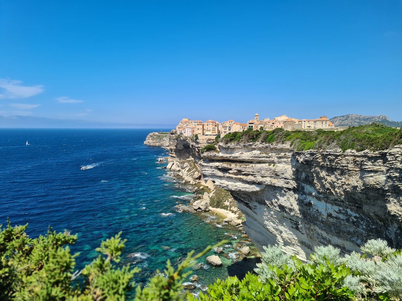 Bright coastal panorama of Bonifacio, Corsica: turquoise sea meets dramatic white limestone cliffs, with a hilltop town beneath blue skies.