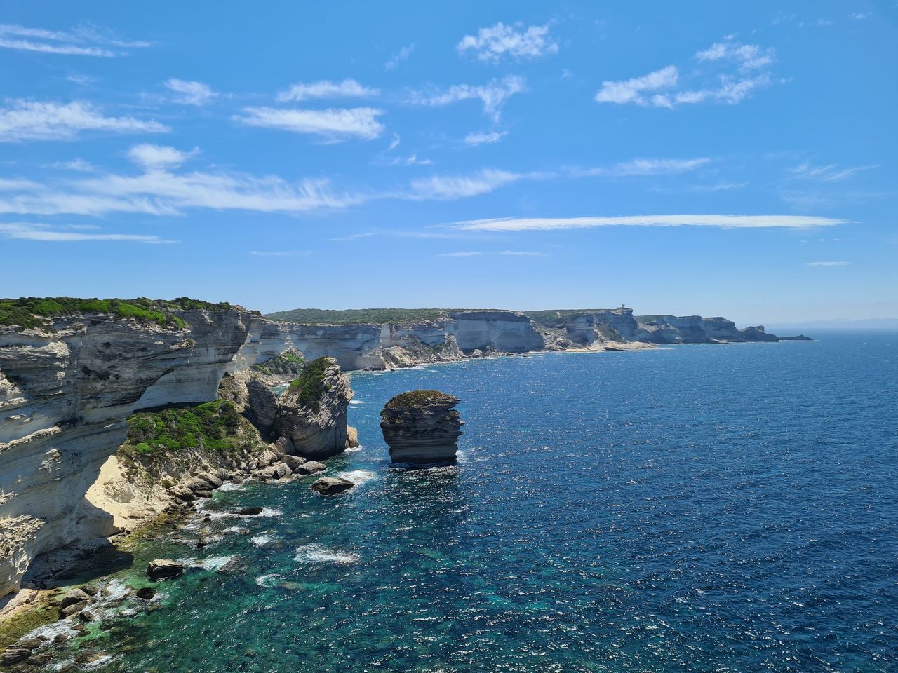 Sunny coastal landscape with rugged limestone cliffs, turquoise waves, and a sea stack beside deep blue ocean under a clear, breezy sky.