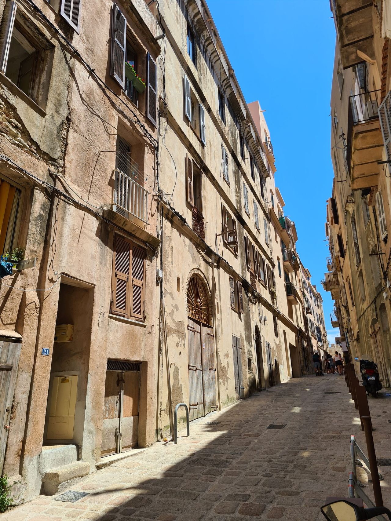 Sunlit narrow cobblestone street lined with weathered stone buildings, shuttered windows, balconies, and a bright blue sky; quiet historic charm.