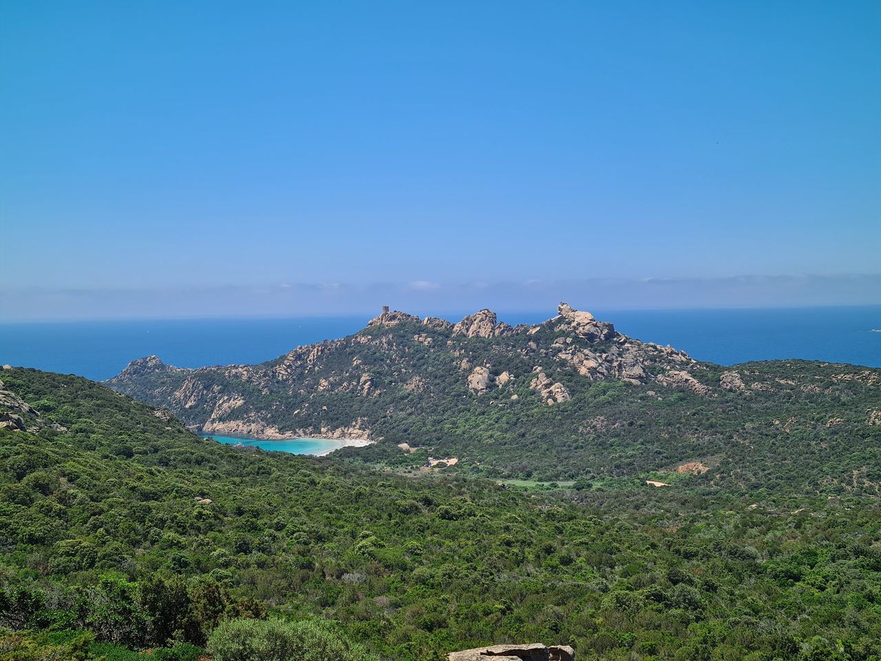 Rugged green coastal mountain ridges rise above turquoise coves and a deep blue sea beneath a clear sky, creating a serene landscape.