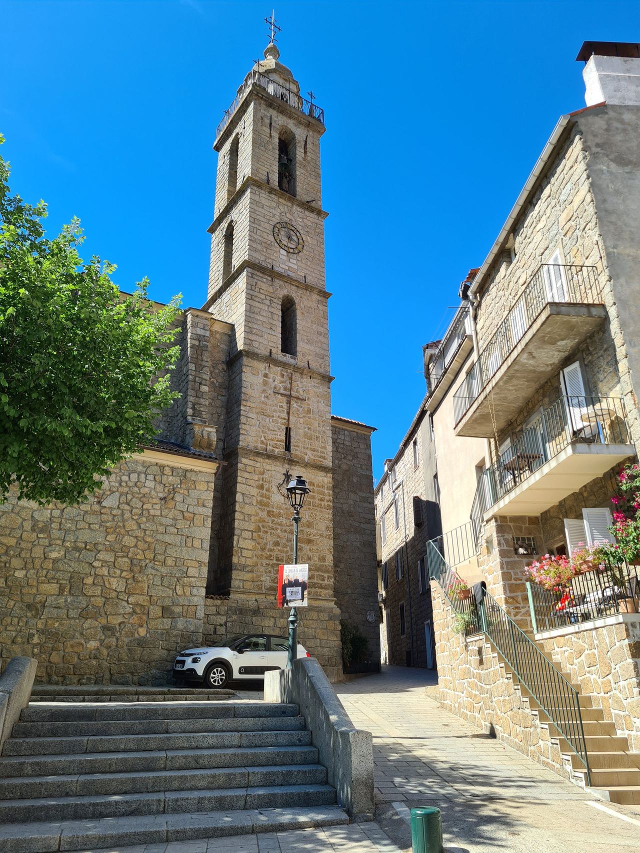 Sunlit historic stone village with a church bell tower and clock, broad steps, flower-filled balconies, leafy tree, and vivid blue sky.