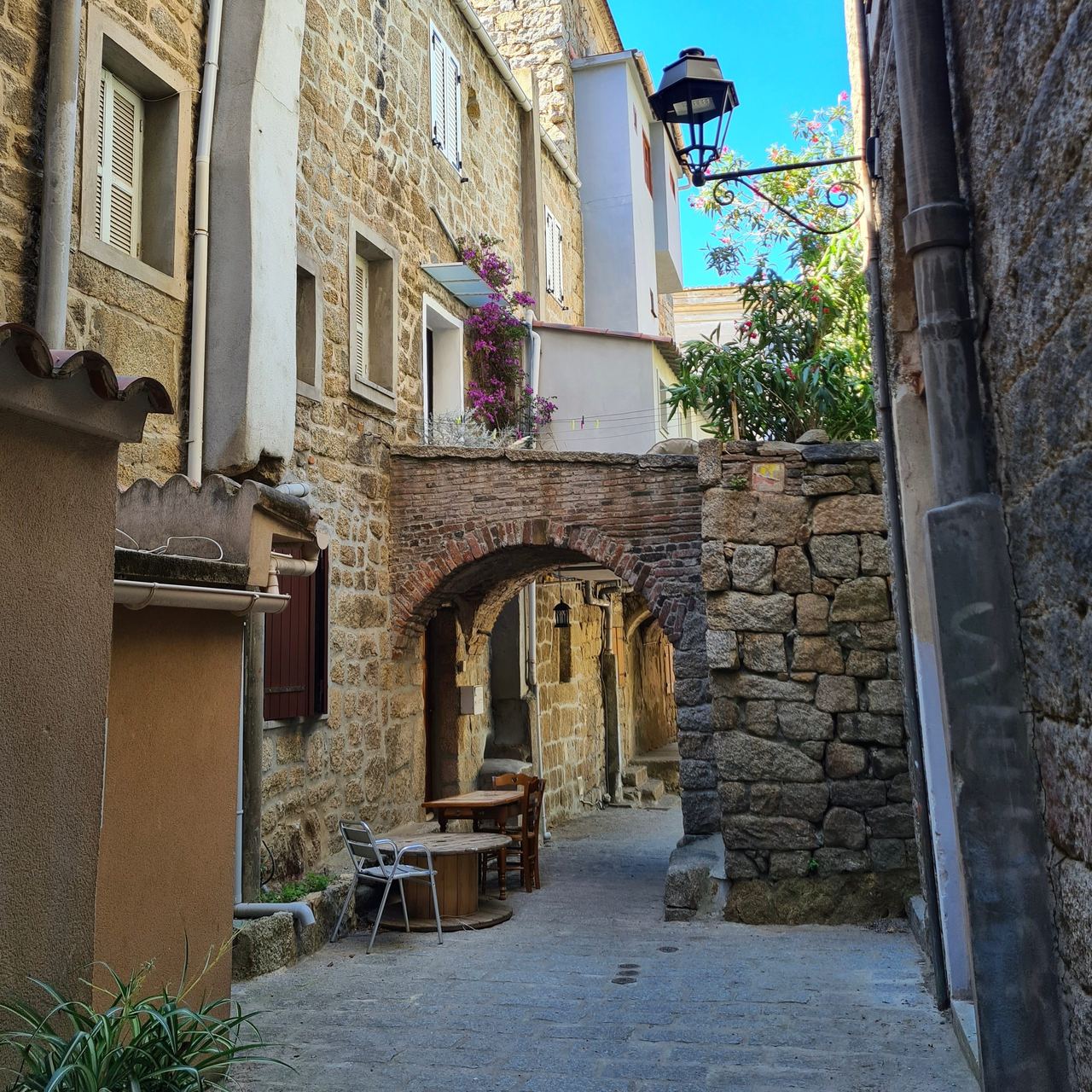 Narrow sunlit stone alley in a historic village, with an arched passage, lantern, flower-covered walls, and a small café table.