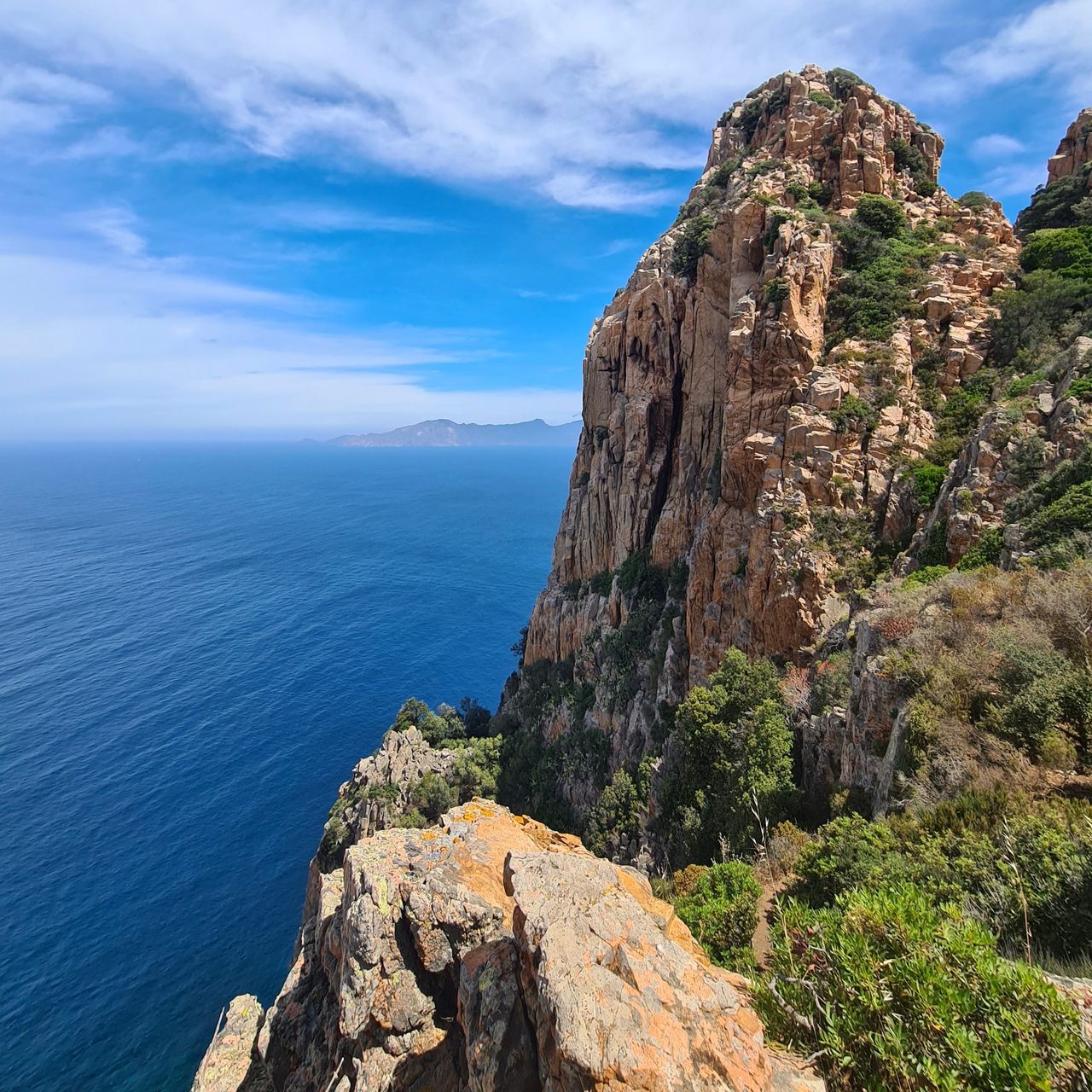 Dramatic coastal sea cliff rises above deep blue water, with rugged orange rock, green shrubs, distant mountains, and a bright, serene sky.