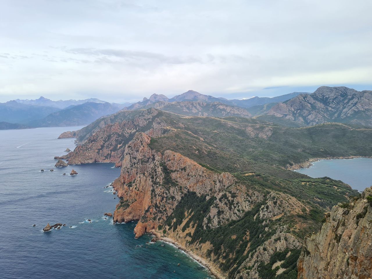 Sweeping coastal mountain landscape with rugged red cliffs, turquoise coves, deep blue sea, and hazy peaks beneath a cloudy sky.