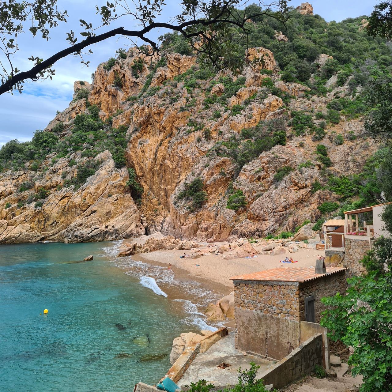 Turquoise cove beneath rugged orange cliffs, with a sandy beach, swimmers, stone buildings, and leafy trees creating a tranquil coastal scene.