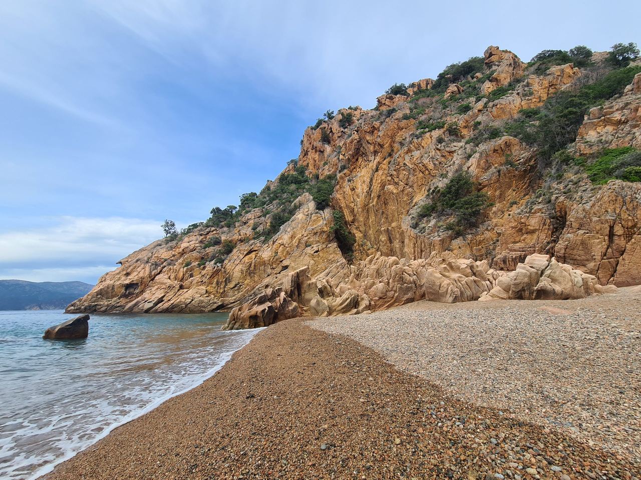 Secluded pebble beach along a rugged coast, beneath towering ochre cliffs, with calm blue water, distant hills, and a peaceful sky.