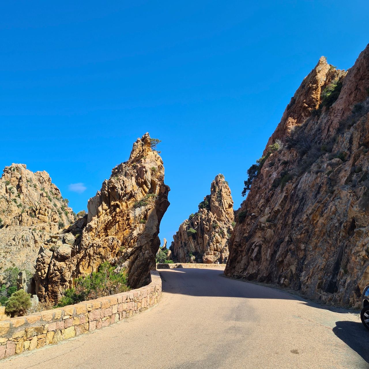 Sunny mountain road winding between dramatic jagged granite cliffs, with a low stone wall, sparse greenery, and vivid blue sky.