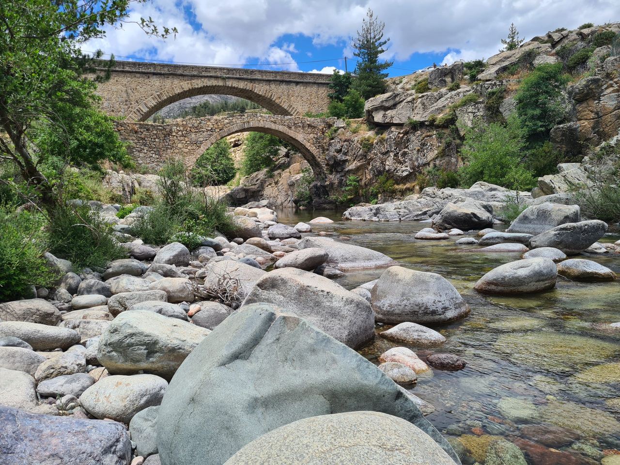 Tranquil mountain stream winds among smooth boulders beneath a historic double-arch stone bridge, framed by trees, cliffs, and blue sky.