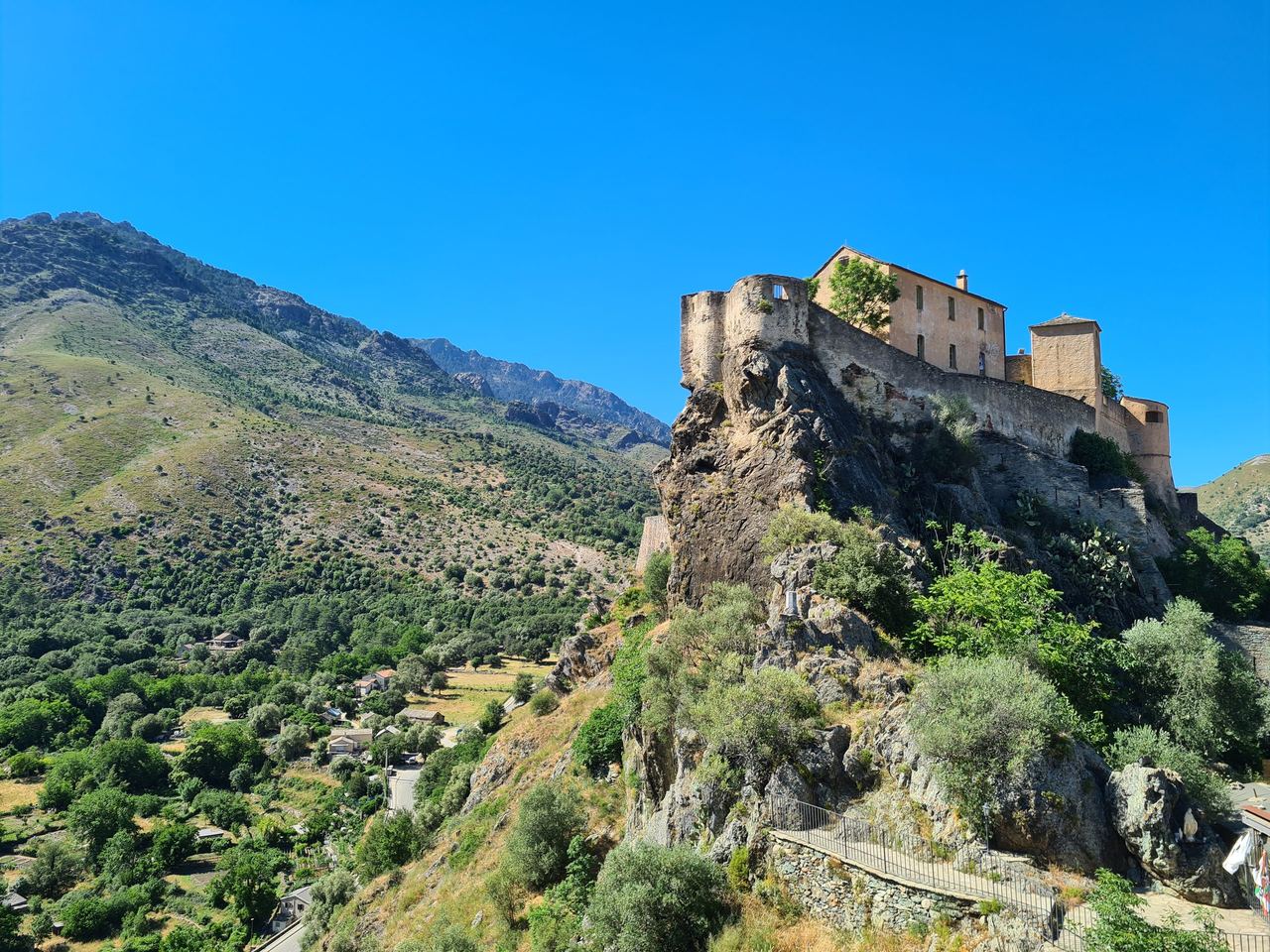 Sunlit medieval stone castle perched on a rugged cliff above a green valley, with mountain slopes, trees, and a vivid blue sky.