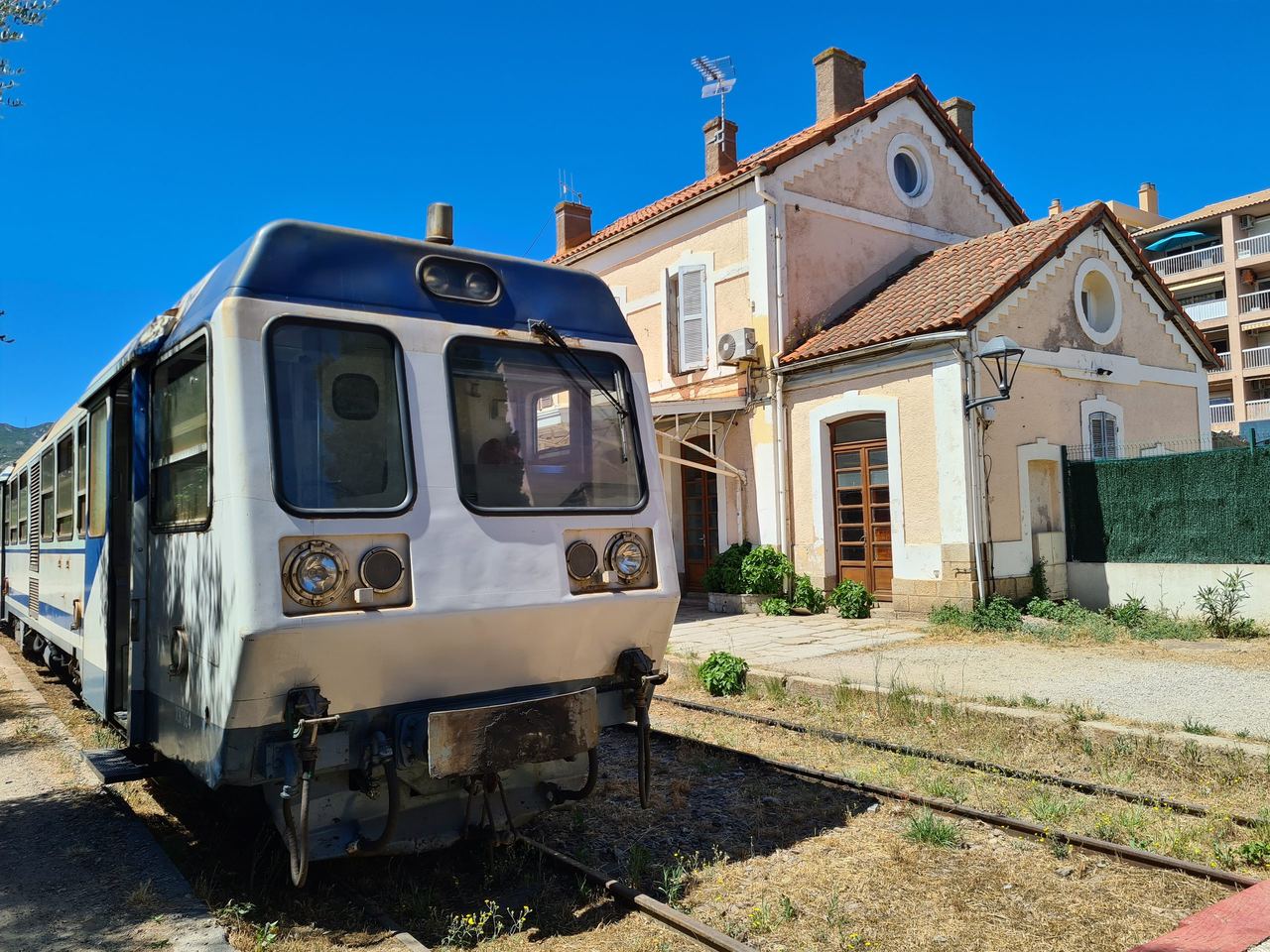Vintage railcar stopped beside a charming Mediterranean station, with terracotta roofs, blue sky, tracks, and bright sunlight creating a nostalgic mood.
