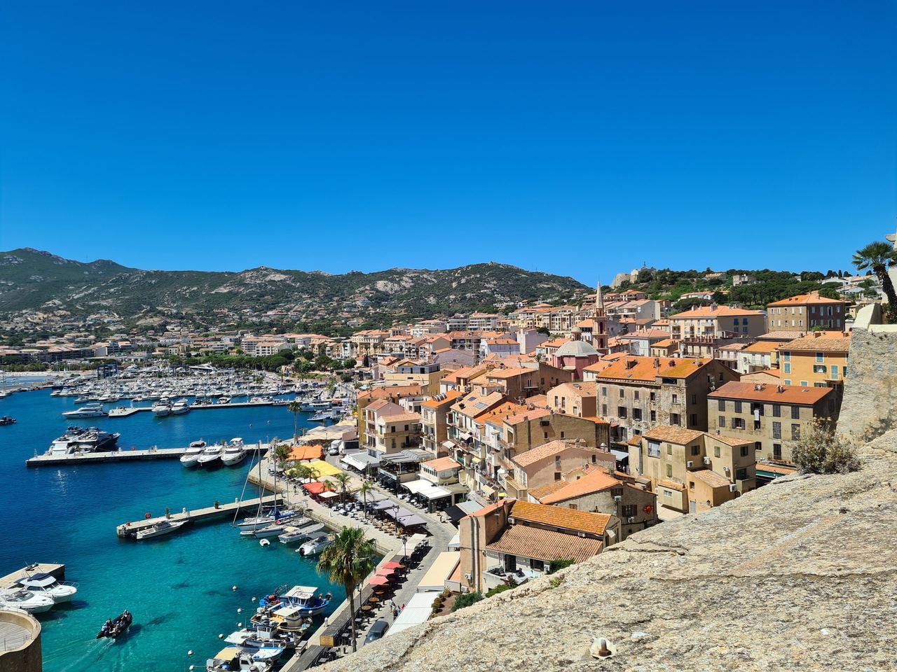 Panoramic view of a sunlit Mediterranean harbor town, with terracotta rooftops, turquoise water, boats, and rugged green hills under blue sky.