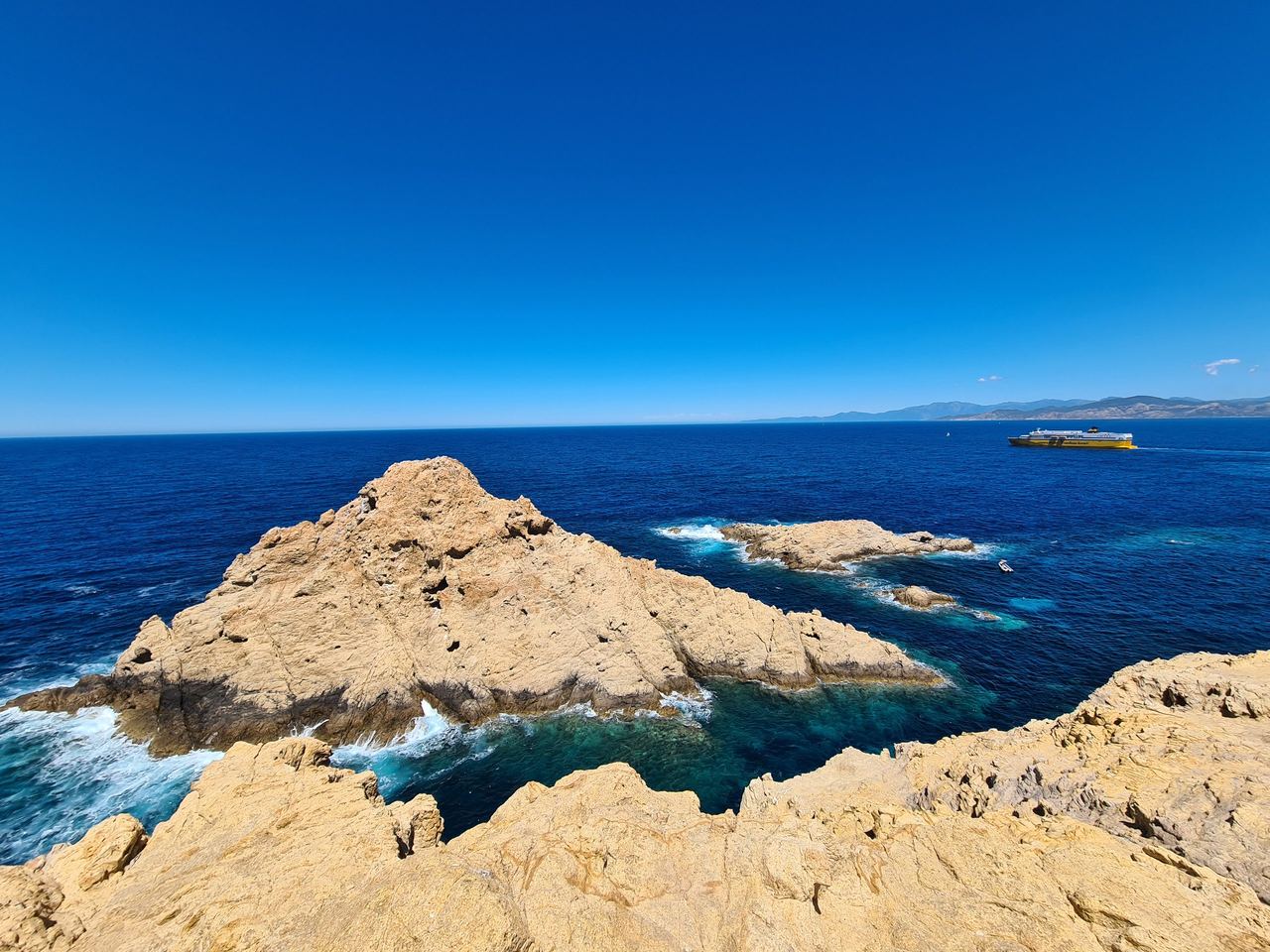 Sunlit golden rock formations rise from deep blue Mediterranean waters, with foamy waves, a distant boat, and a clear, serene sky.