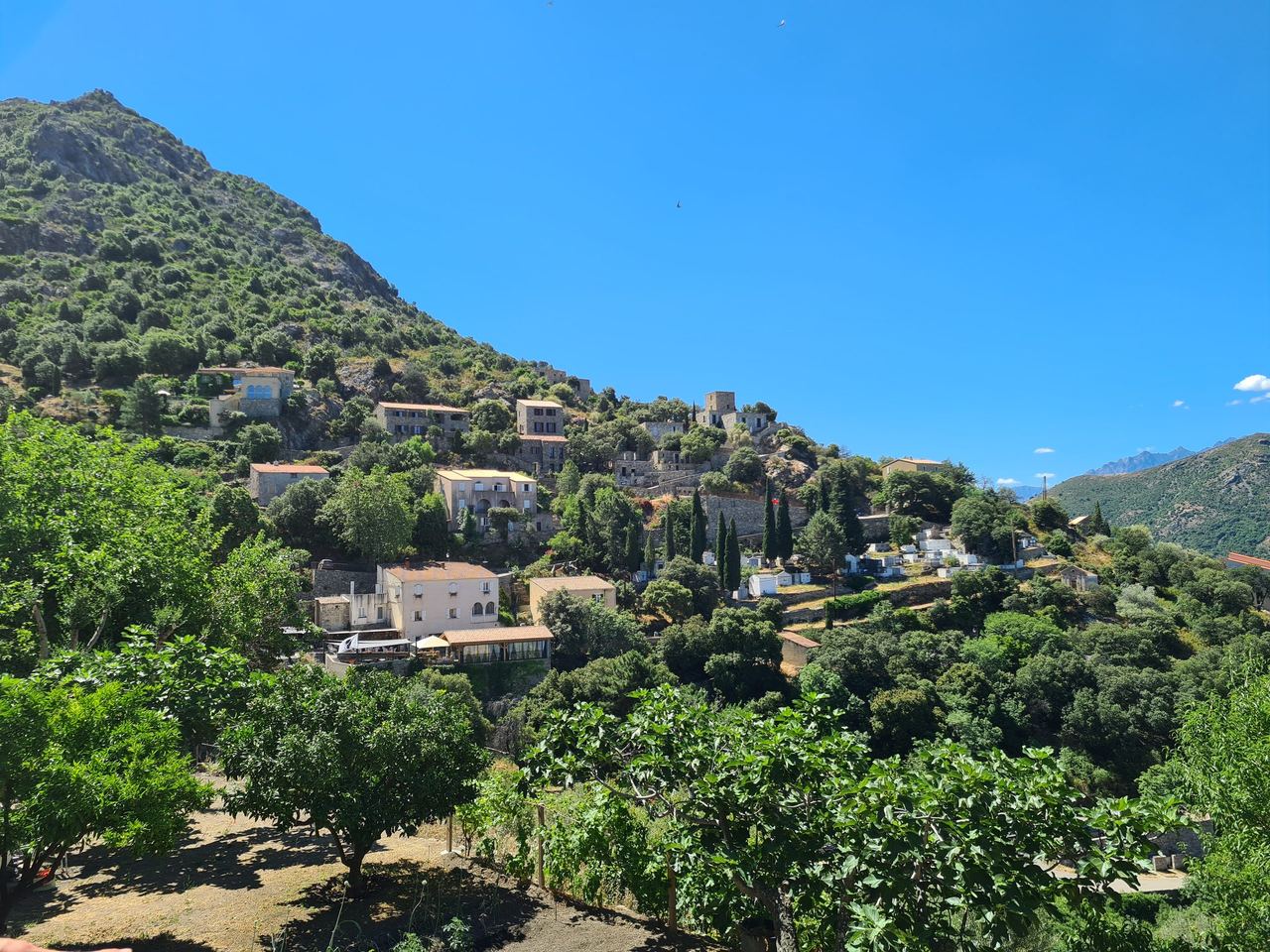 Sunlit hillside village with terracotta-roofed homes, lush orchards, and rugged green mountains beneath a clear blue sky; serene Mediterranean scene.