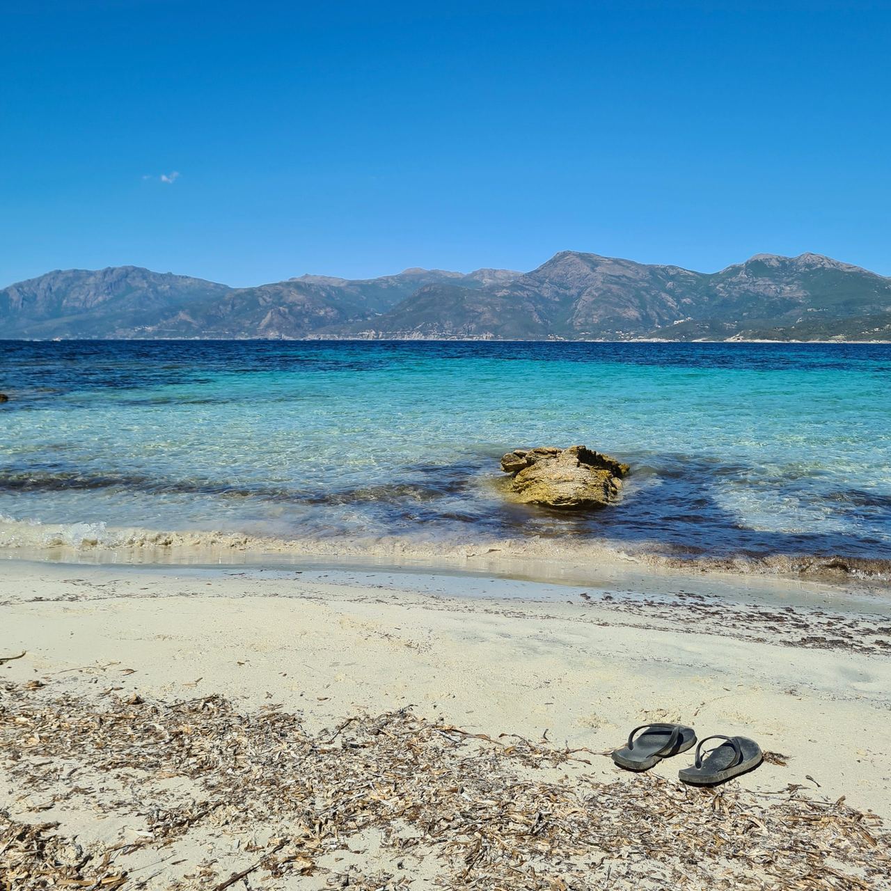 Serene tropical beach with turquoise shallows, a rocky islet, distant rugged mountains, and black flip-flops on sunlit sand under blue skies.