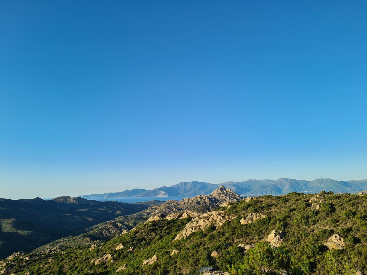 Sunlit mountain landscape with rocky, shrub-covered ridges in the foreground, distant blue peaks, and a vast clear sky creating a serene mood.