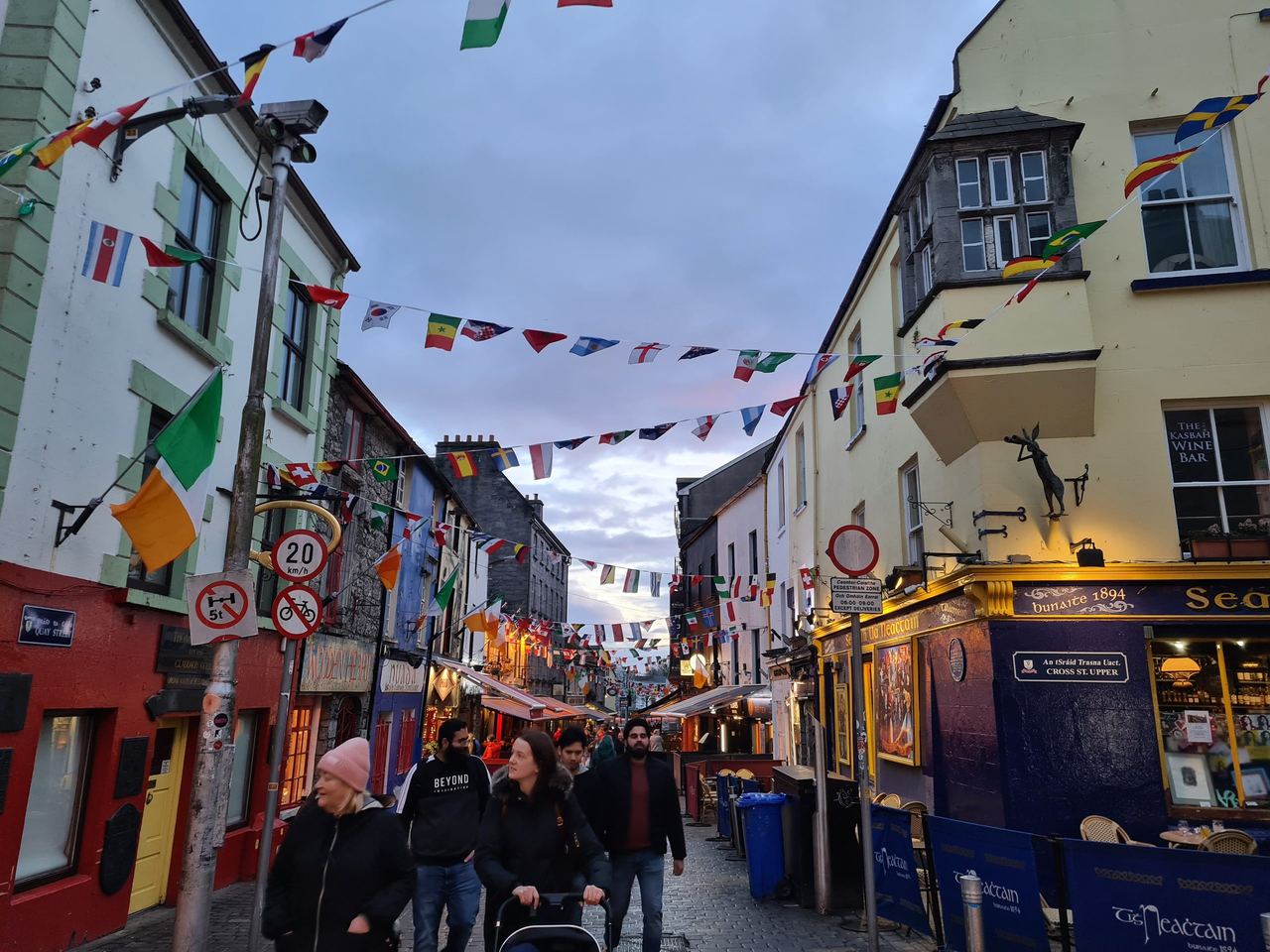Pedestrian street of a town with people walking around under hanging Irish flags on a cloudy evening