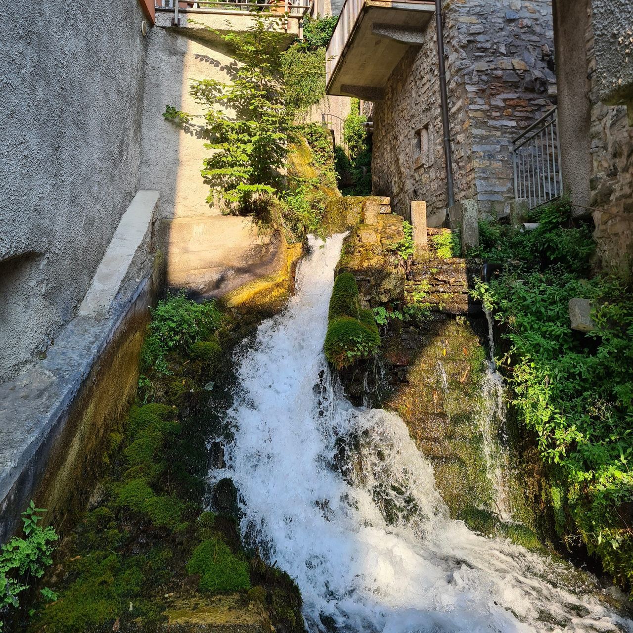 A narrow village waterfall rushes over mossy stone beneath old buildings, blending rustic architecture, greenery, and a serene natural mood.