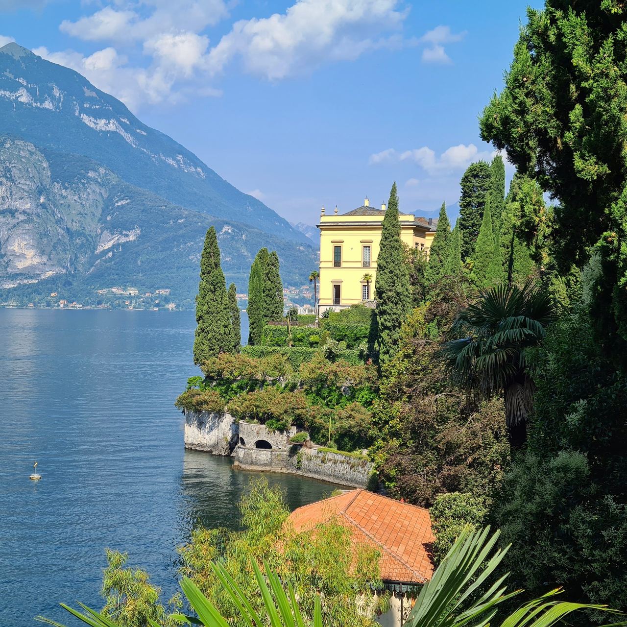 Sunny Lake Como villa on a lush, cypress-lined peninsula, with terracotta roofs, blue water, and rugged mountains beneath a clear sky.