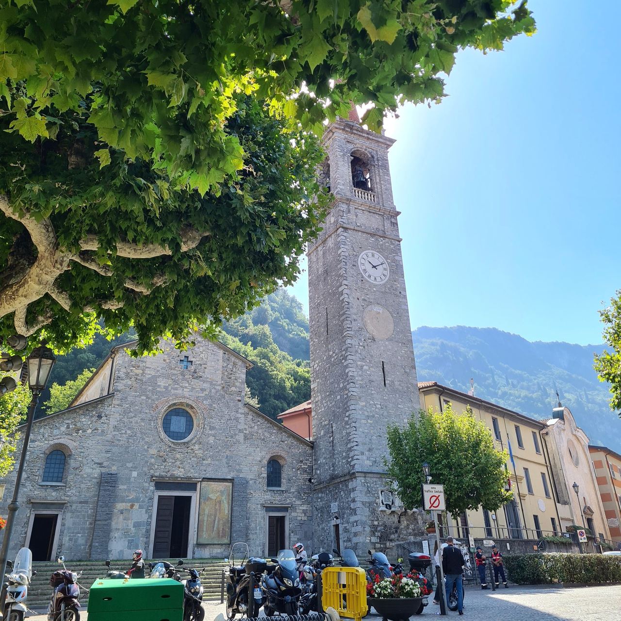 Historic stone church and clock tower beneath leafy trees, with a mountain backdrop, village street, scooters, and pedestrians in sunlight.
