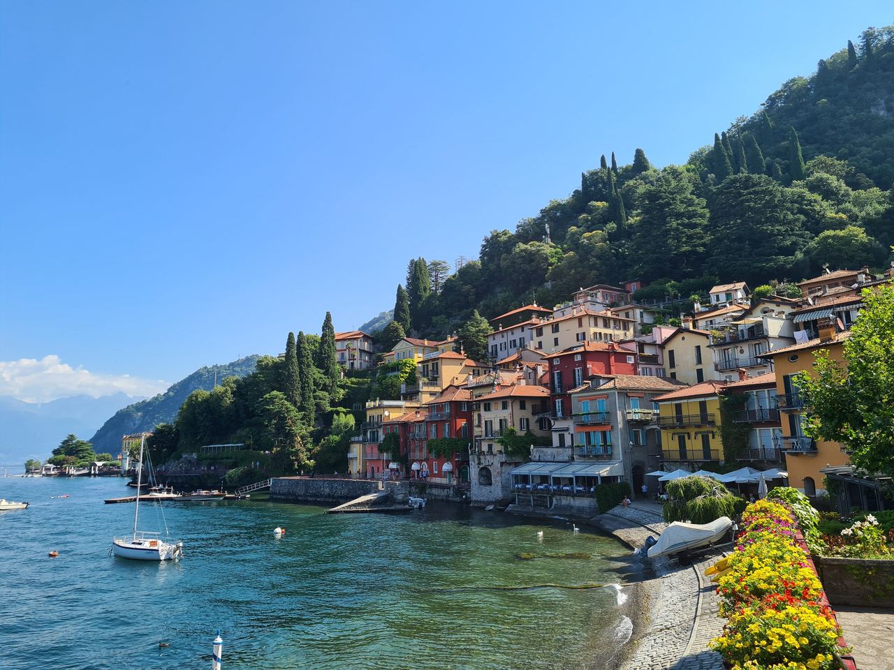 Sunny Italian lakeside village with colorful hillside homes, cypress trees, boats, and turquoise water beneath a clear blue sky; tranquil mood.