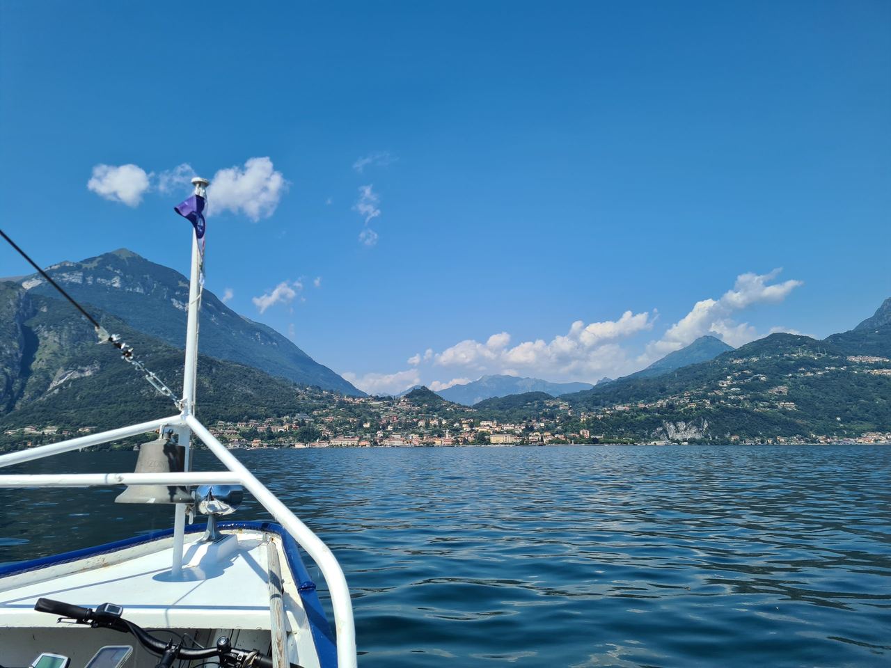 View from a boat across a sparkling alpine lake toward green, village-dotted mountains beneath a vivid blue sky; peaceful summer scene.