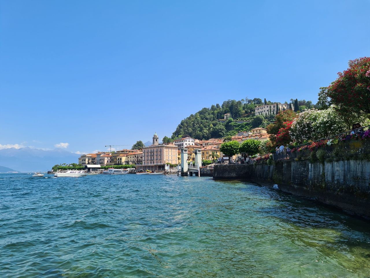 Scenic lakeside village with colorful waterfront buildings, lush hillside homes, and a stone promenade beside sparkling blue-green water under clear sky.
