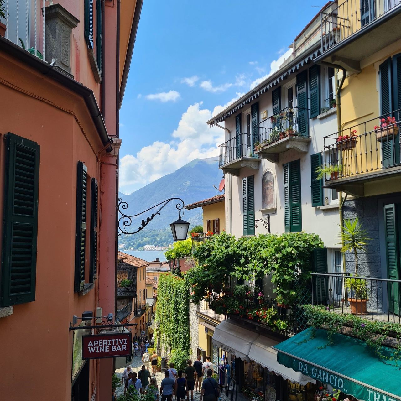 Narrow Italian lakeside street with colorful buildings, flower-filled balconies, a wine bar sign, pedestrians, and mountains under a sunny sky.