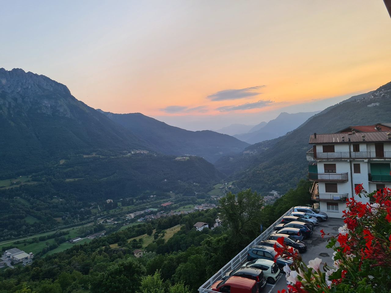 Sunset illuminates a mountain valley and winding village below, with a hillside apartment, parked cars, and bright red flowers in foreground.