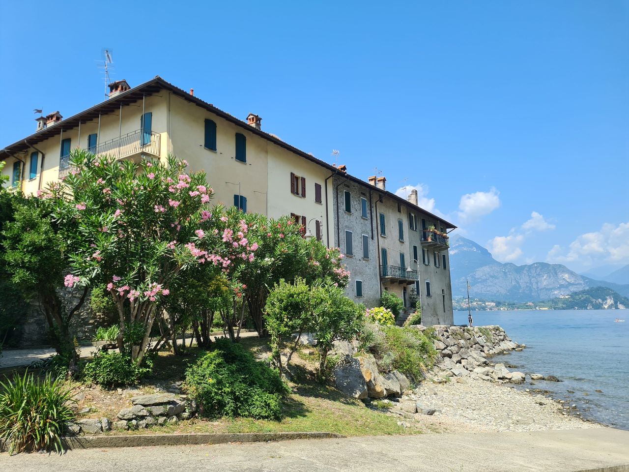 Historic lakeside stone villa with pink oleanders, rocky shore, and blue mountain lake beneath a clear sky, evoking a peaceful Italian retreat.