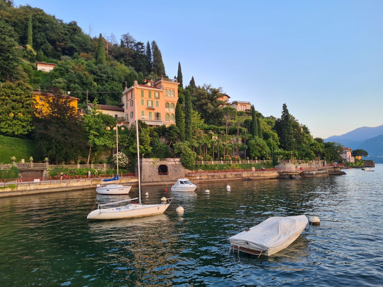Lakeside Italian villa surrounded by lush gardens, sailboats and moored boats on calm water, with distant mountains under a blue sky.