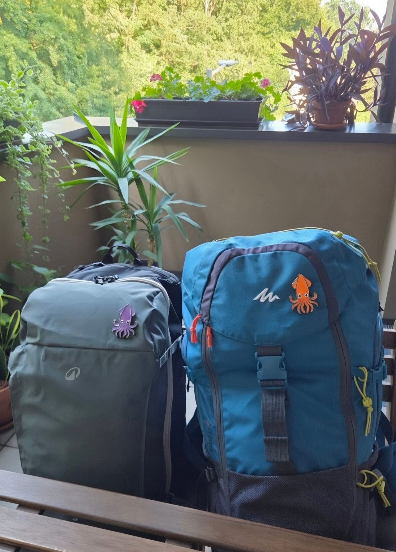 Two travel backpacks—gray and blue with playful patches—sit on a balcony bench amid lush green plants, creating a calm, outdoorsy scene.