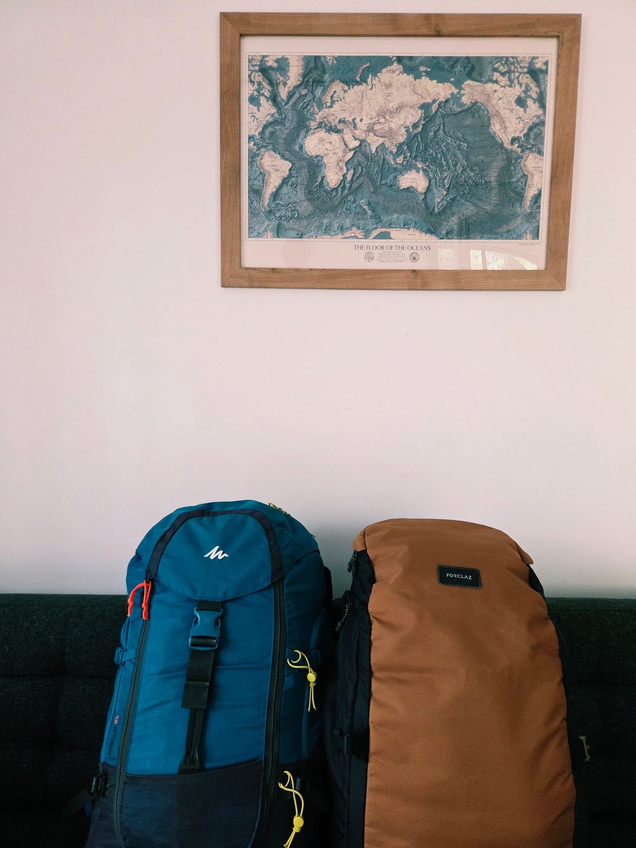 Two hiking backpacks, one blue and one brown, rest on a dark sofa beneath a framed vintage world map, evoking travel and adventure.