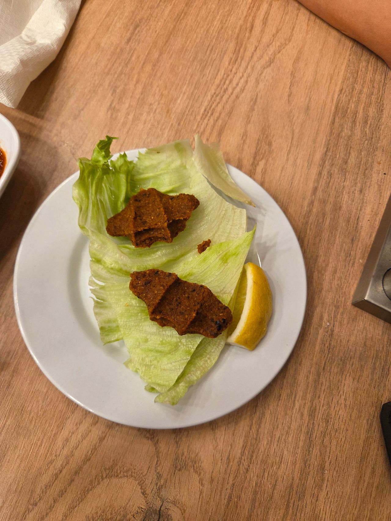 A white plate holds crisp lettuce leaves topped with browned köfte pieces and a lemon wedge on a wooden table, suggesting a simple, casual meal.