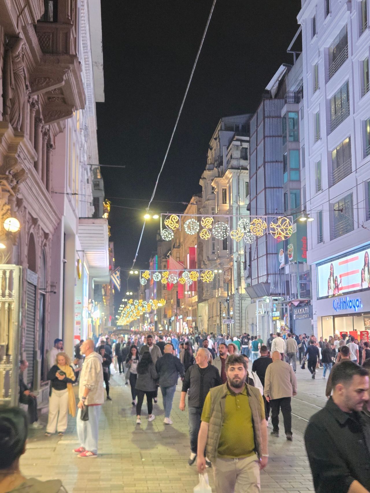 A crowded pedestrian street in Istanbul glows beneath festive string lights at night, with historic and modern buildings framing a lively, bustling scene.