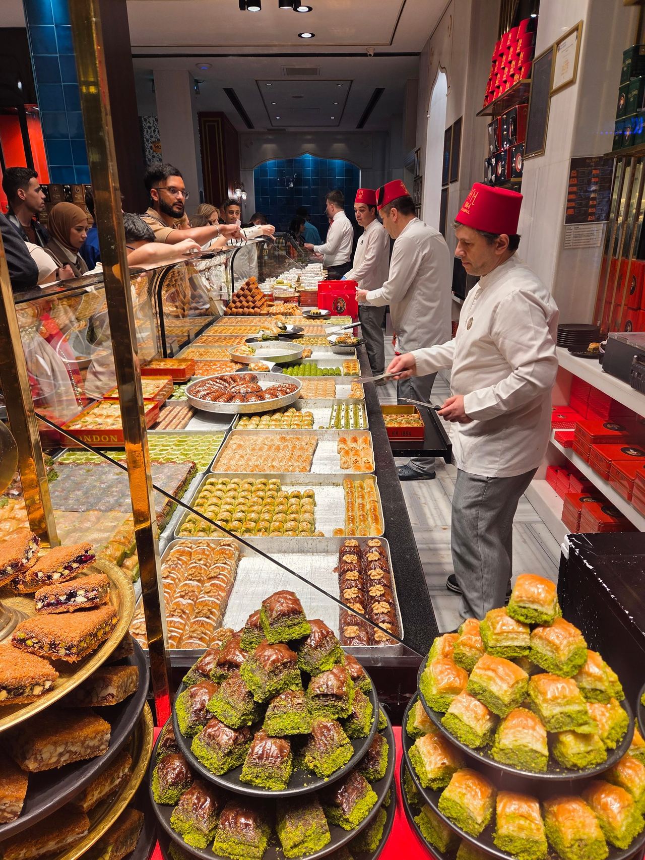 A bustling Middle Eastern sweets shop, with chefs in white uniforms and red hats serving customers behind glass displays of baklava and pastries.