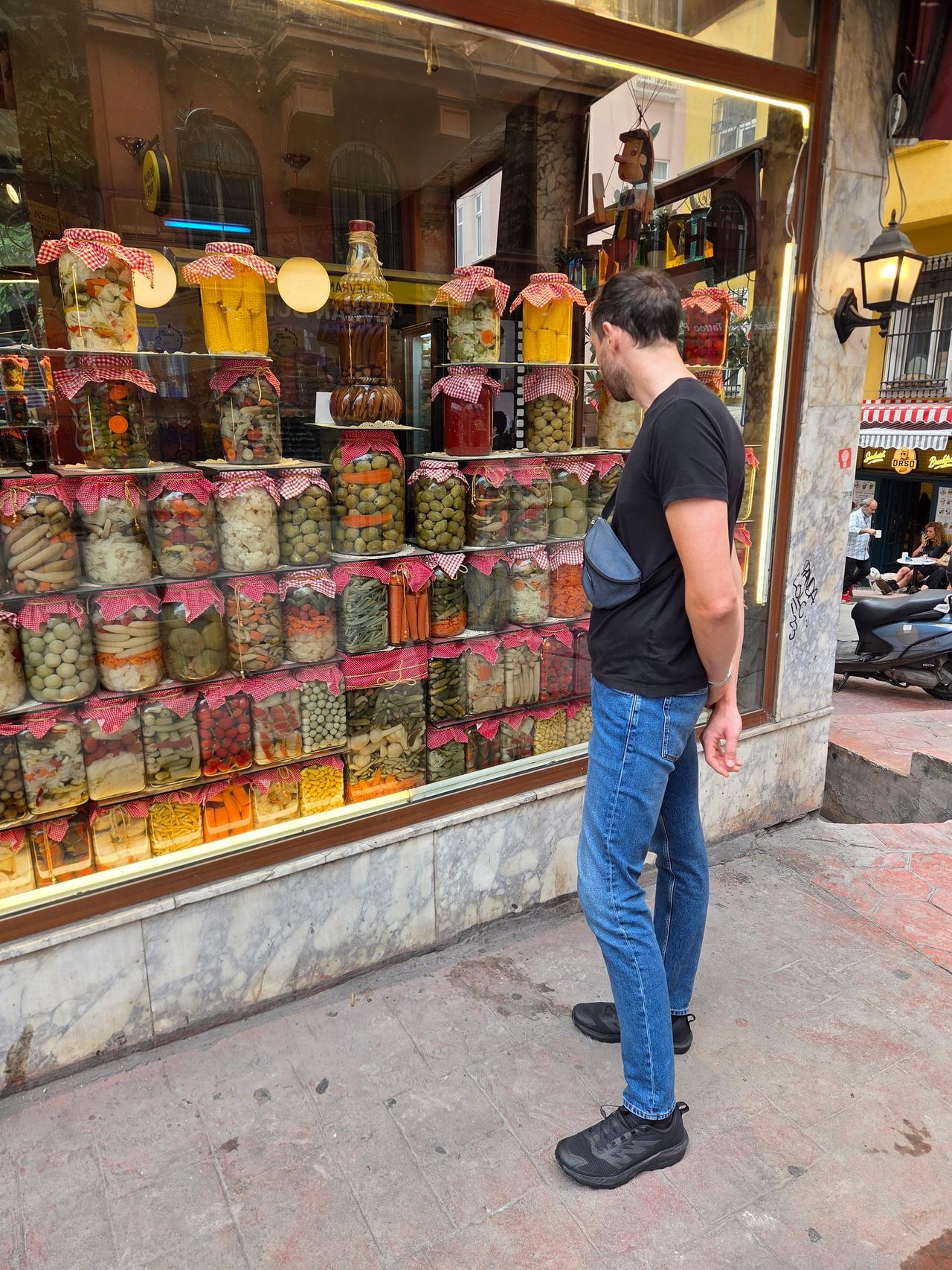A man studies a shop window packed with colorful jars of pickled vegetables, olives, and preserves