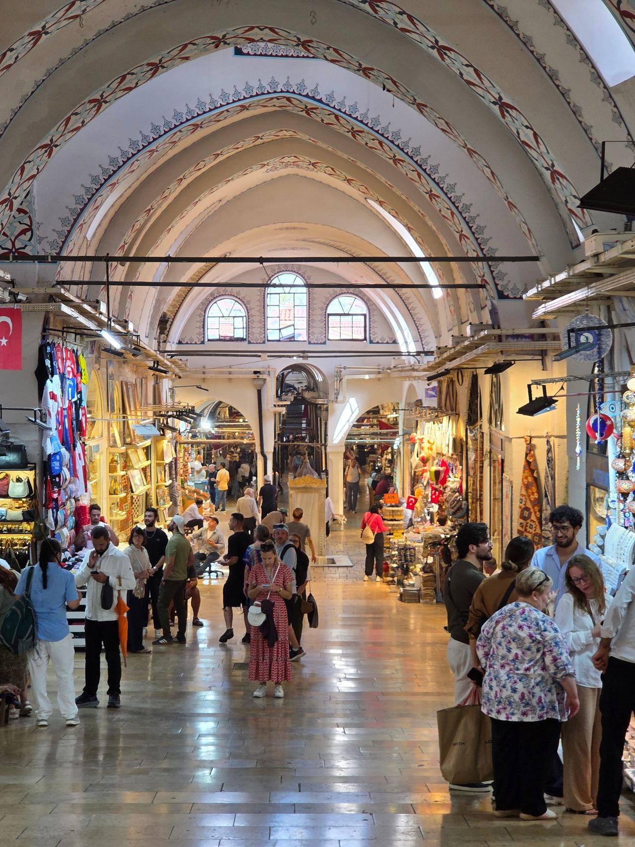 Istanbul’s Grand Bazaar bustles with shoppers among colorful stalls, beneath ornate vaulted arches and stained-glass windows in a historic hall.