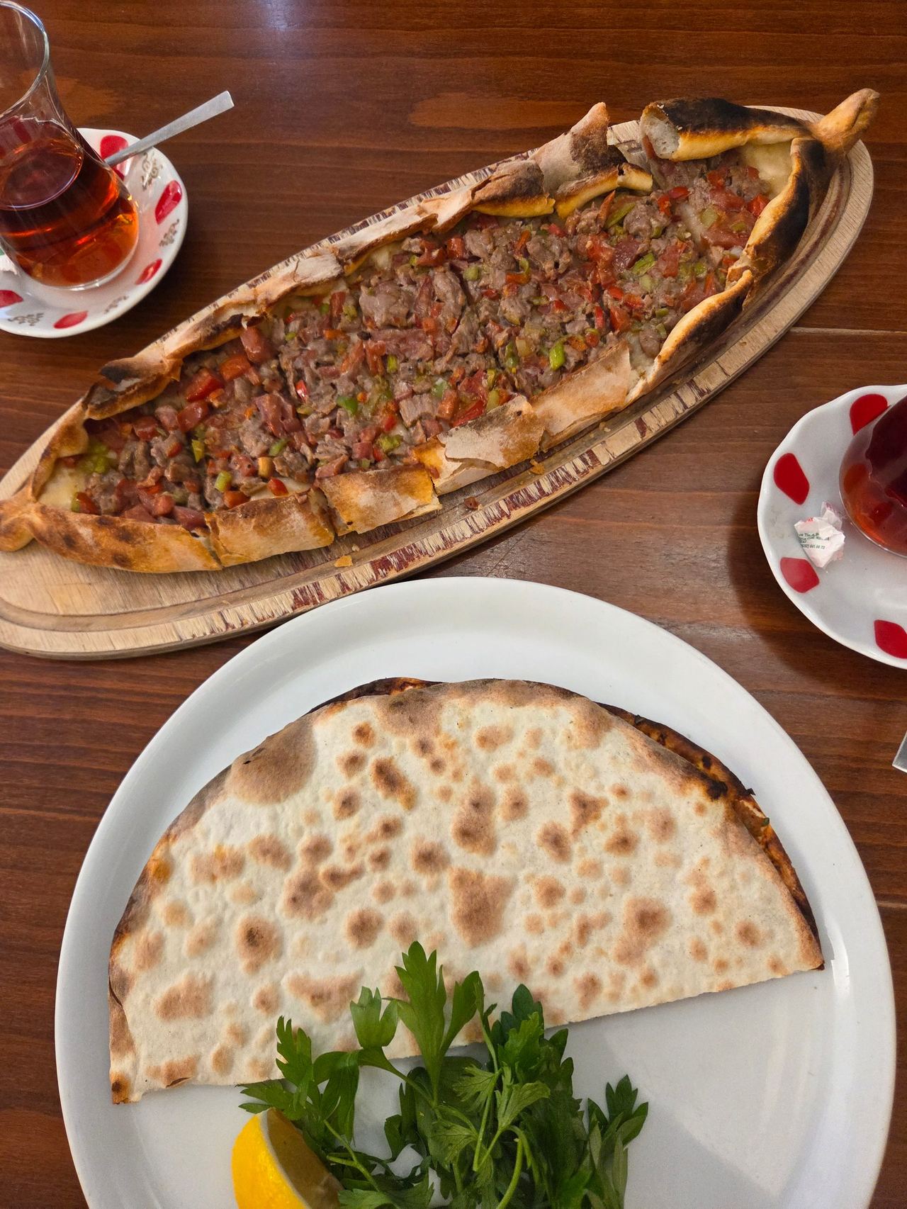 Overhead view of Turkish pide topped with minced meat and vegetables beside flatbread with parsley and lemon, plus glasses of tea on a wooden table.