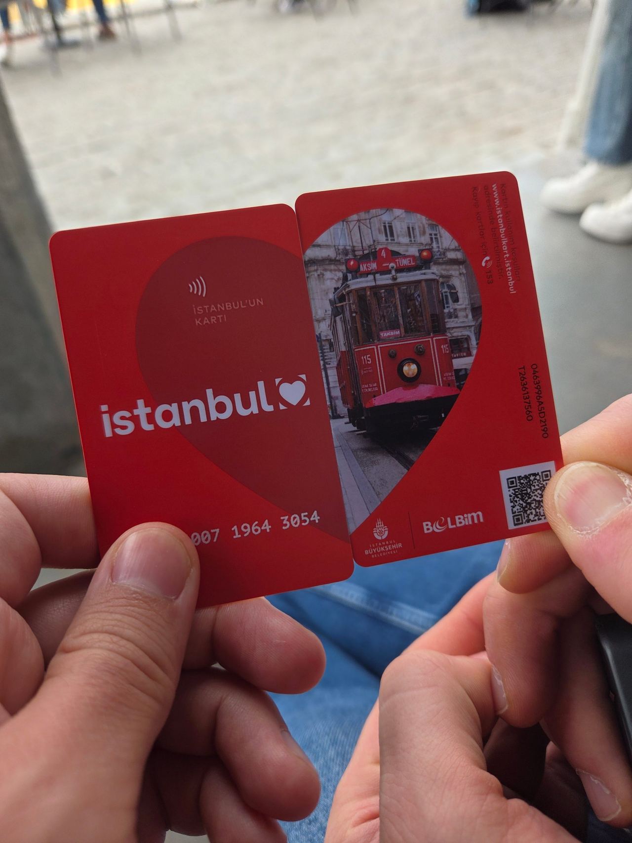 Hands hold two red Istanbul transit cards, one showing a vintage tram and QR code, against a blurred city street.