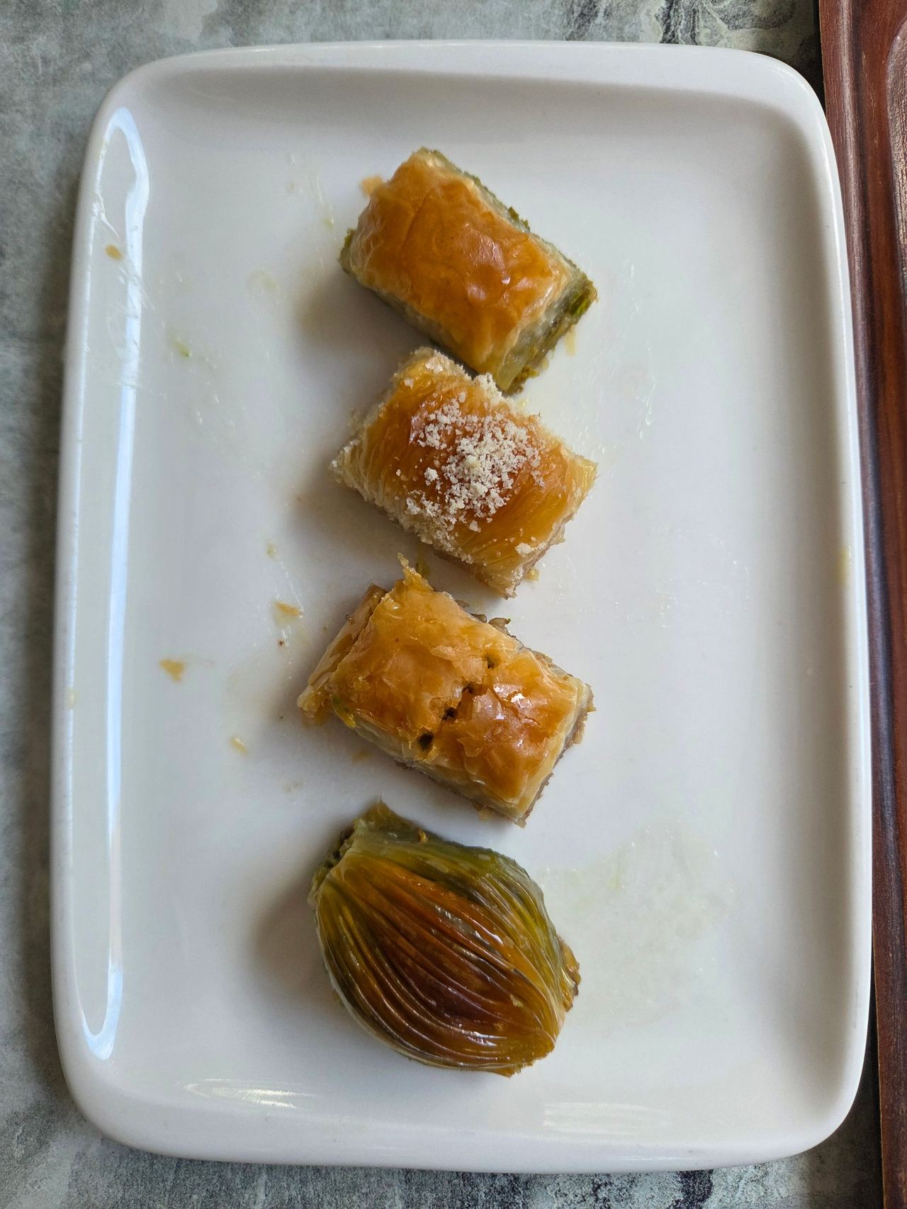 Four golden pieces of syrup-soaked baklava, including a layered pistachio-filled piece, arranged vertically on a white plate; a simple, appetizing presentation.
