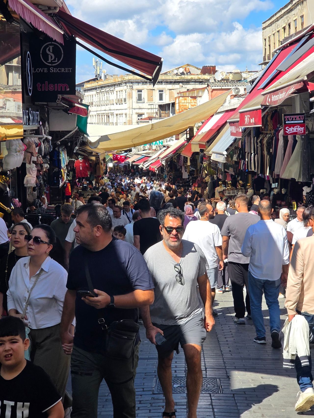A crowded outdoor market fills a narrow street, with shoppers beneath colorful awnings and shop signs in bright sunlight.