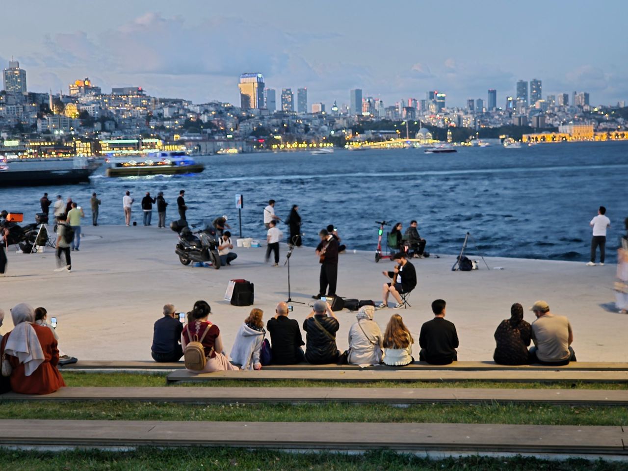 At dusk, at Sarayburnu Park, people sit and stroll along at the waterfront, watching boats cross the Bosphorus as a brightly lit city skyline glows behind them.