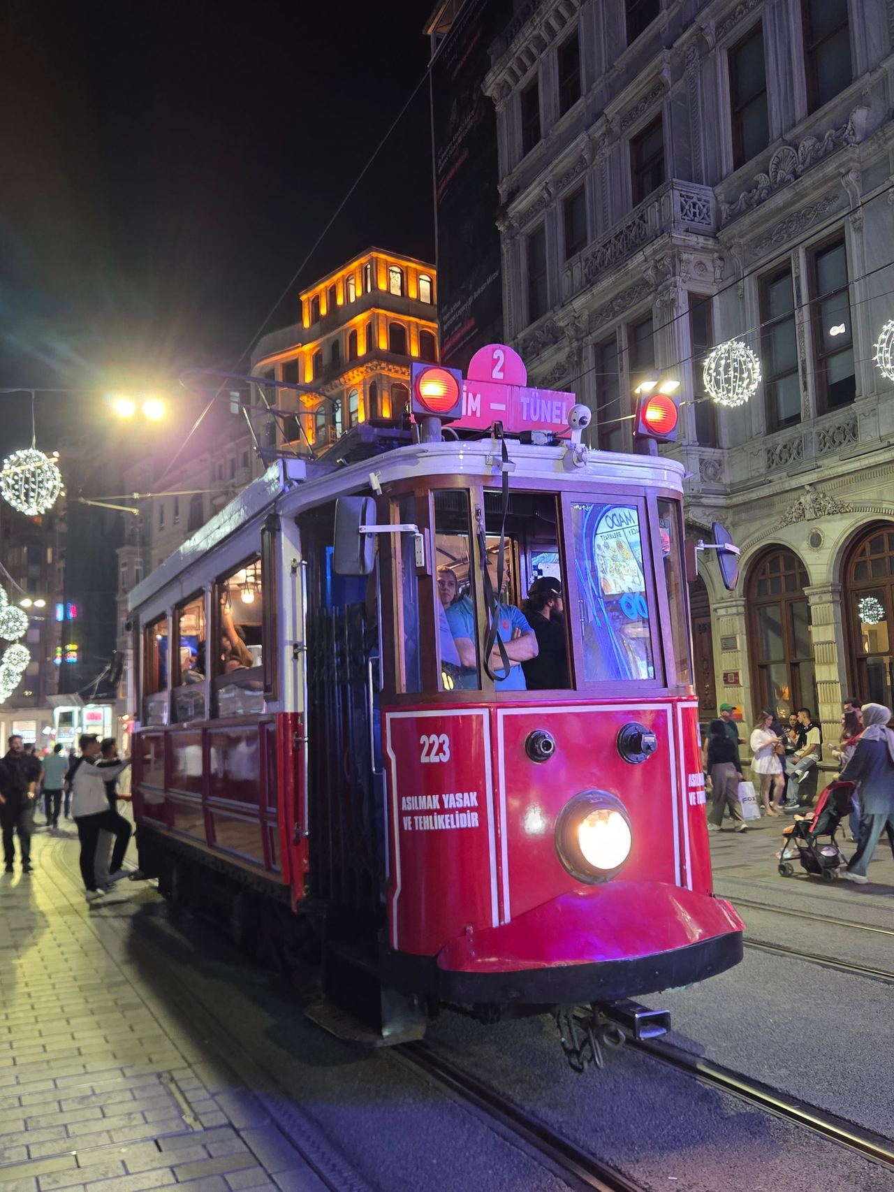 A magenta Istanbul heritage tram approaches along a busy city street at night, its headlight glowing beneath ornate buildings and festive lights.
