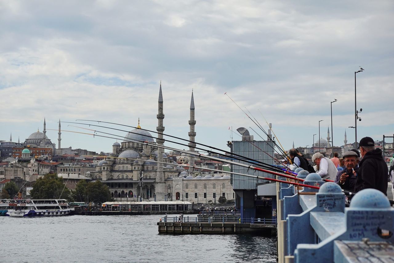 People fish from Istanbul’s Galata Bridge over the Bosphorus, with boats and historic mosques and minarets along a cloudy, busy waterfront.