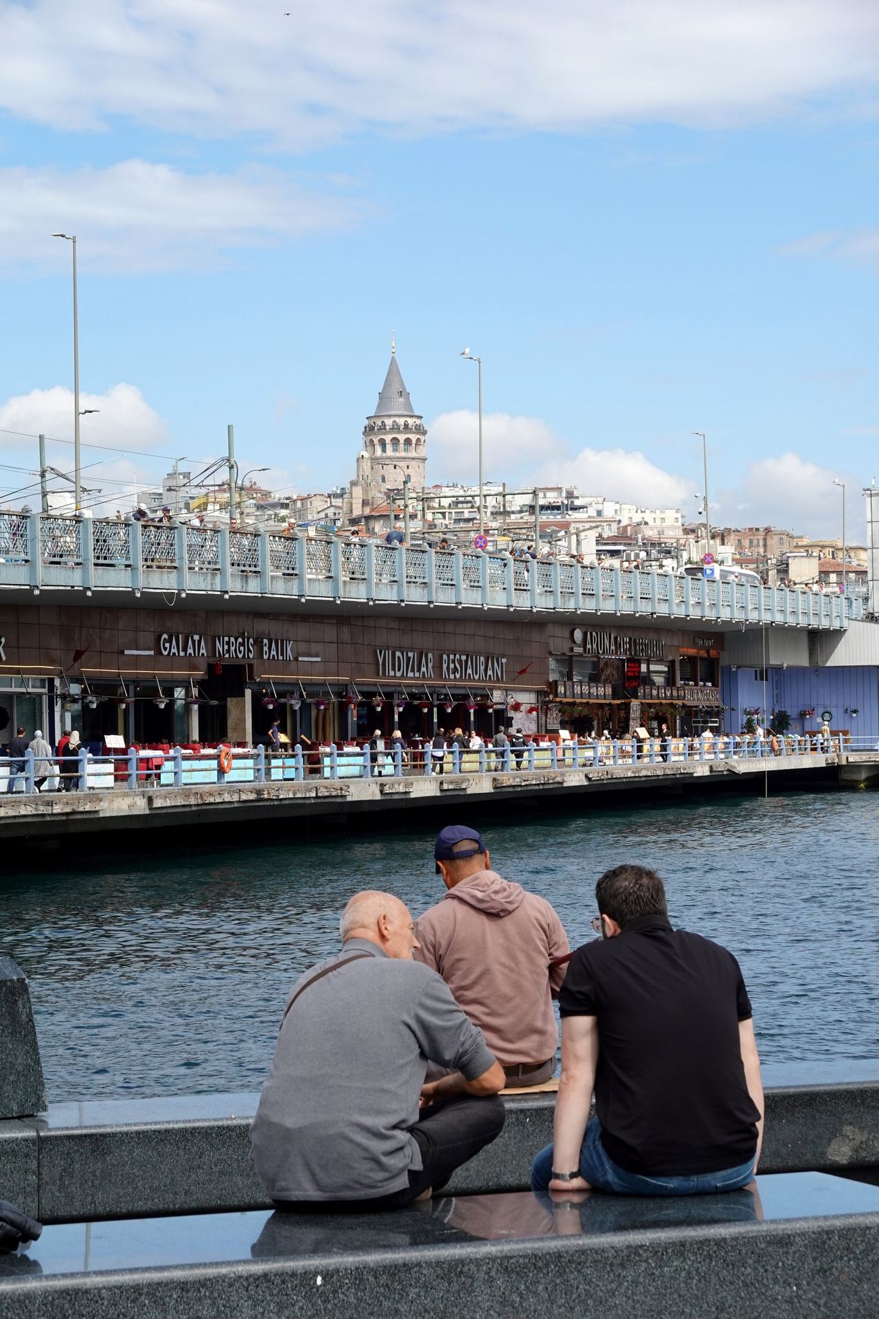 Three men sit along a waterfront railing, gazing across busy Galata Bridge toward Istanbul’s Galata Tower under a bright, partly cloudy sky.