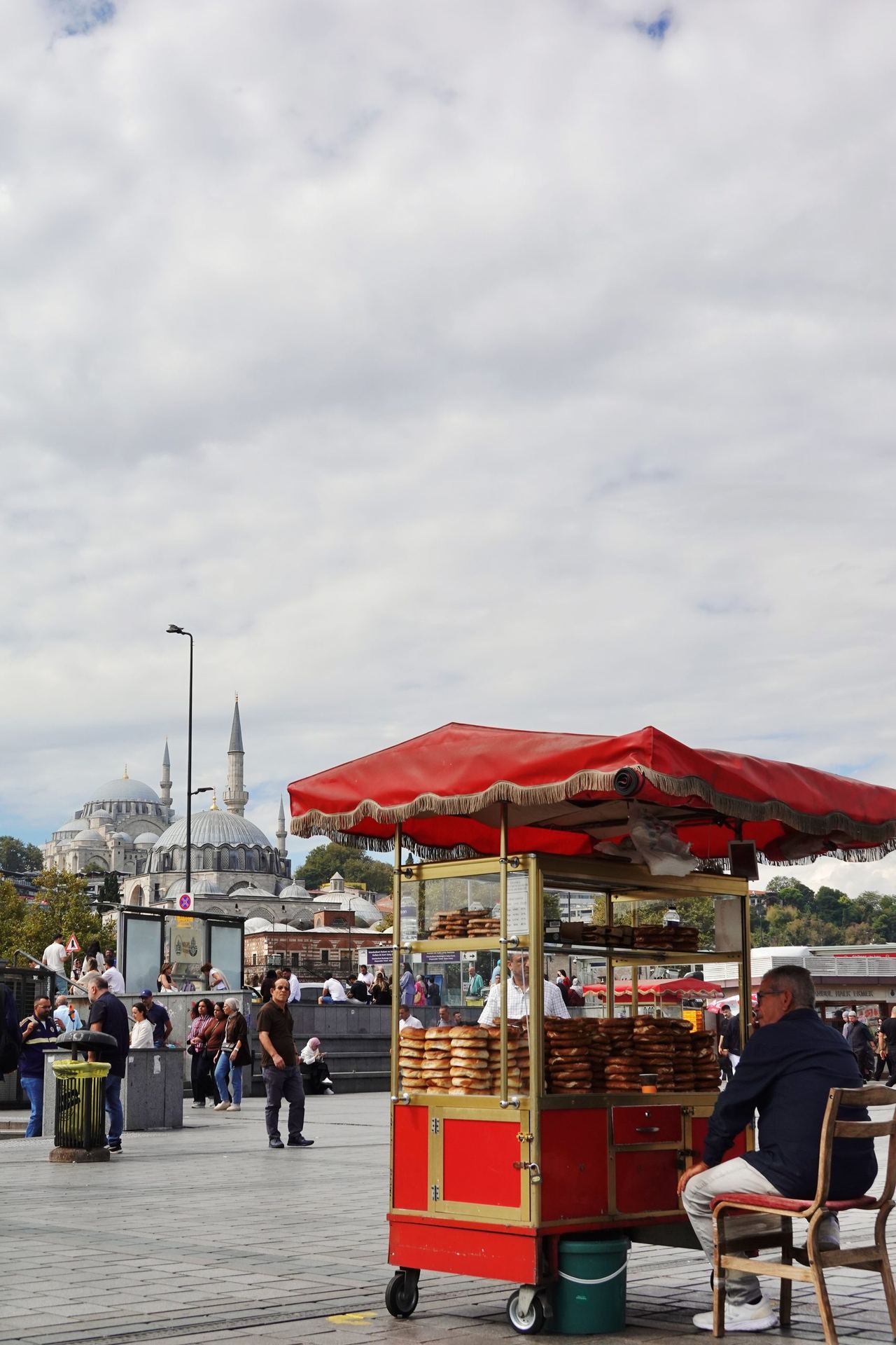 A red-roofed street food cart selling simit stands on a busy Istanbul waterfront, with a seated vendor, crowds and mosque minarets beneath a cloudy sky.