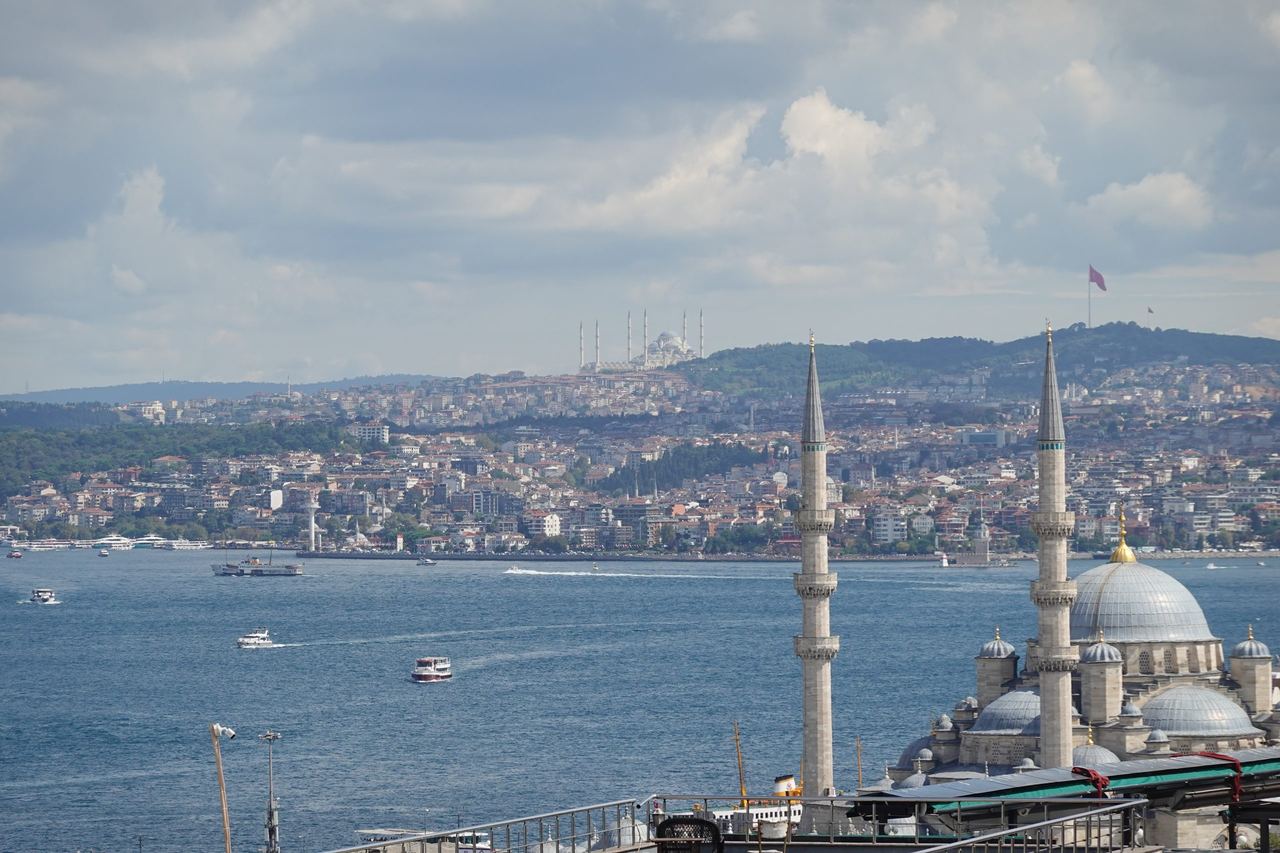 Istanbul waterfront with Yeni Mosque in the foreground, boats crossing the blue Bosphorus, and a dense city beneath cloudy skies.