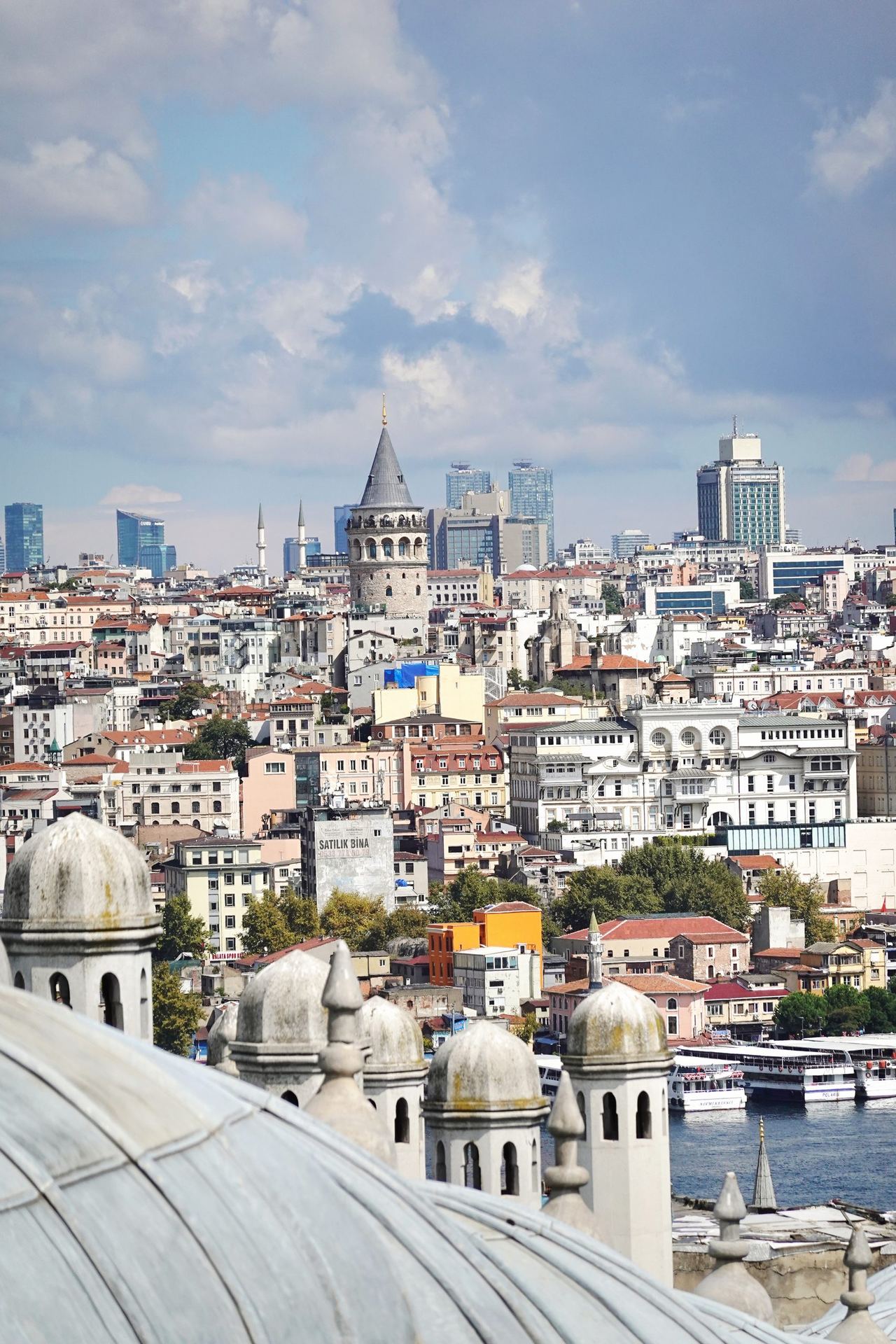 Panoramic view of Istanbul’s Galata Tower rising above dense rooftops, framed by mosque domes and minarets under a bright, calm sky.